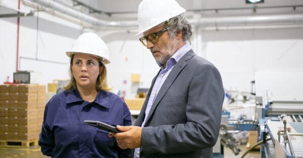 Male engineer and female factory worker in hardhats standing and talking on plant floor, man using tablet. Medium shot. Industrial occupation concept