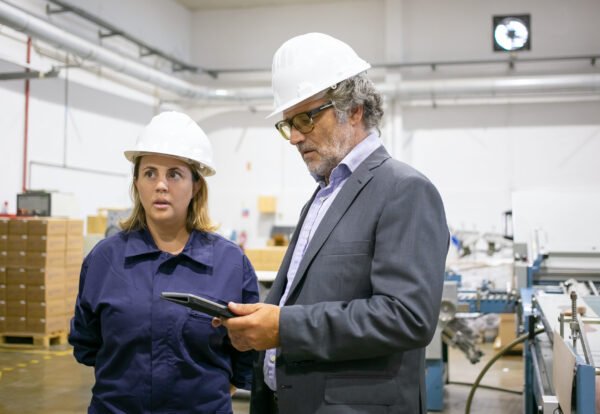 Male engineer and female factory worker in hardhats standing and talking on plant floor, man using tablet. Medium shot. Industrial occupation concept