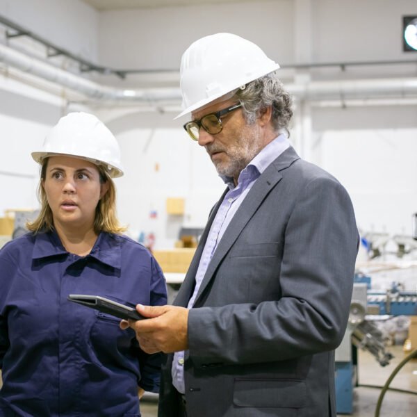 Male engineer and female factory worker in hardhats standing and talking on plant floor, man using tablet. Medium shot. Industrial occupation concept