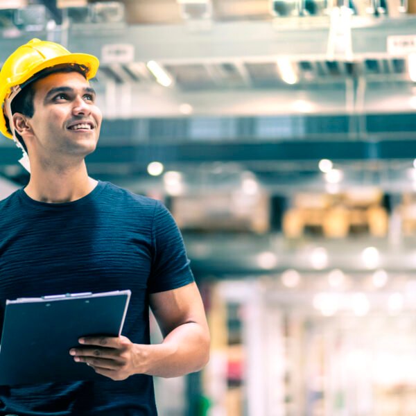 Smart Indian engineer man wearing safety helmet doing stock tick check and cardboard stock product management in factory warehouse background